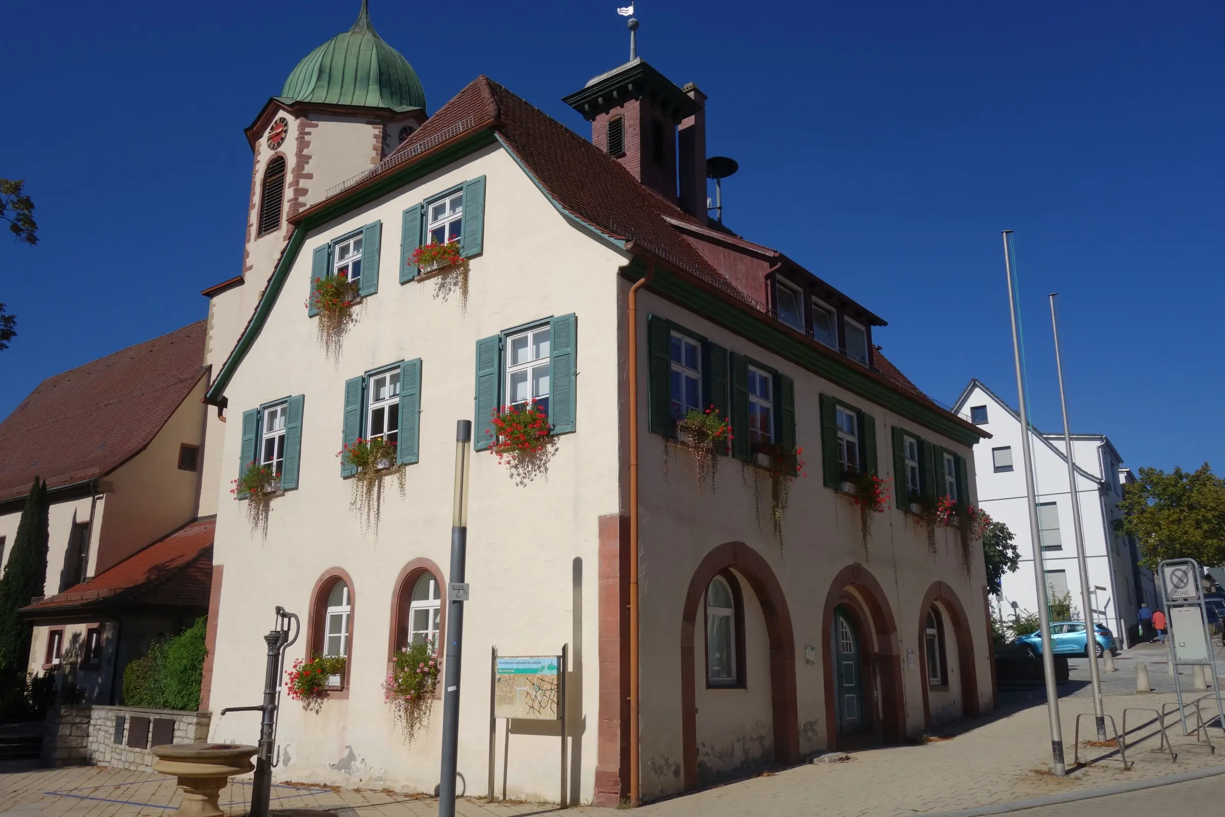 Rathaus Malmsheim mit grünen Fensterläden und Blumenkästen, dahinter der Turm der Germanuskirche, aufgenommen im September 2023